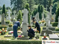 A cemetery. People are cleaning tombstones and placing offerings. There are green trees around.