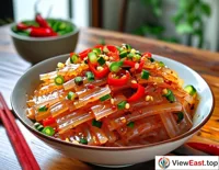 A food blogger is showing homemade Liang Pi in front of the camera, with seasonings and shredded cucumber laid out beside it.